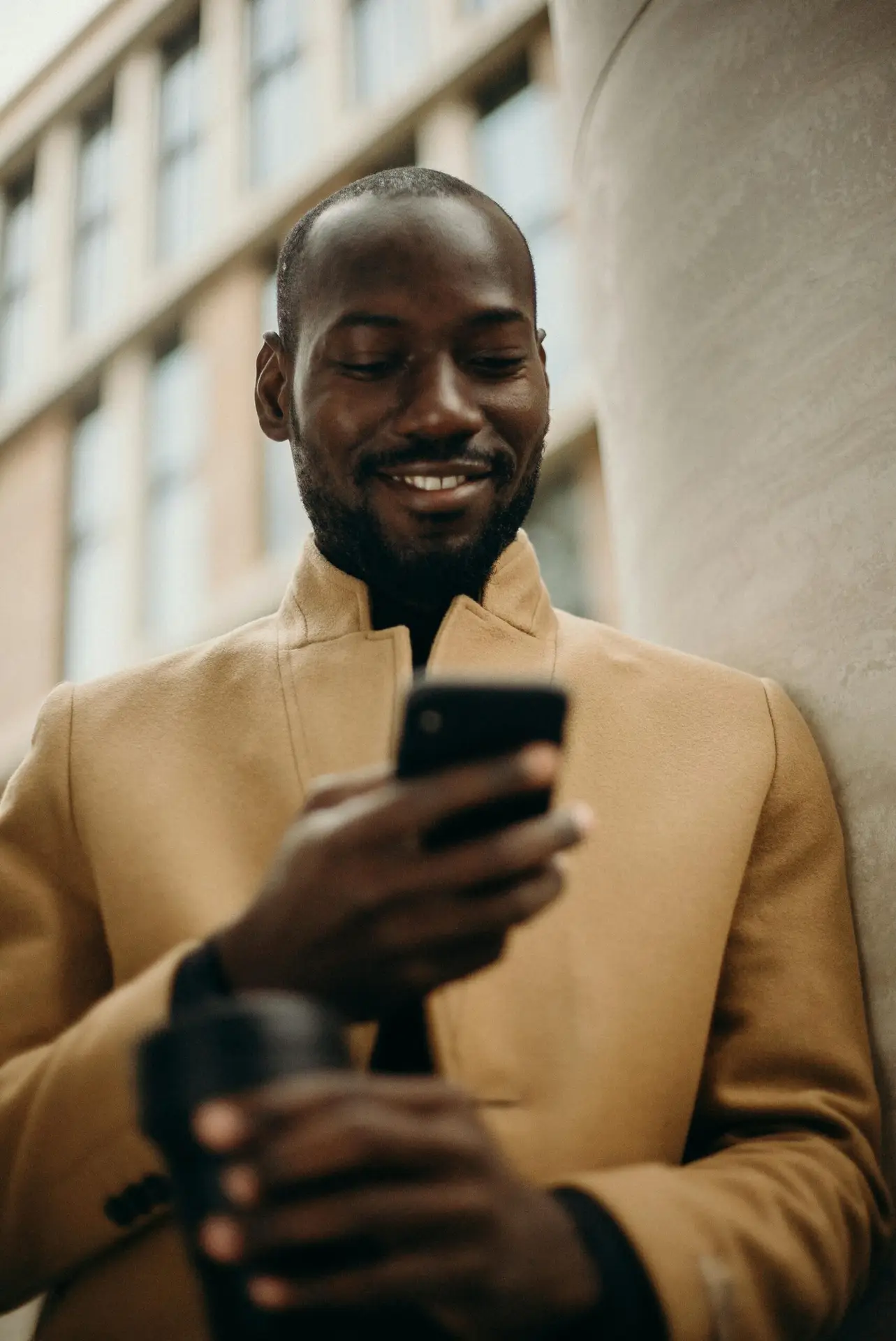 Offerings A cheerful man in a stylish coat using his smartphone and holding coffee outside.